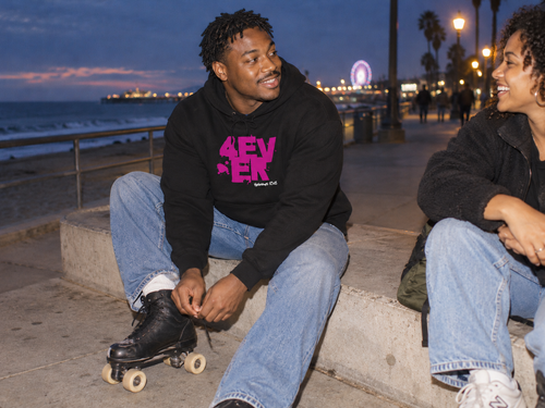 lifestyle mockup of a young adult fastening roller skates with a friend on a boardwalk at dusk, wearing the black 4 Ever hoodie with its magenta front artwork visible.