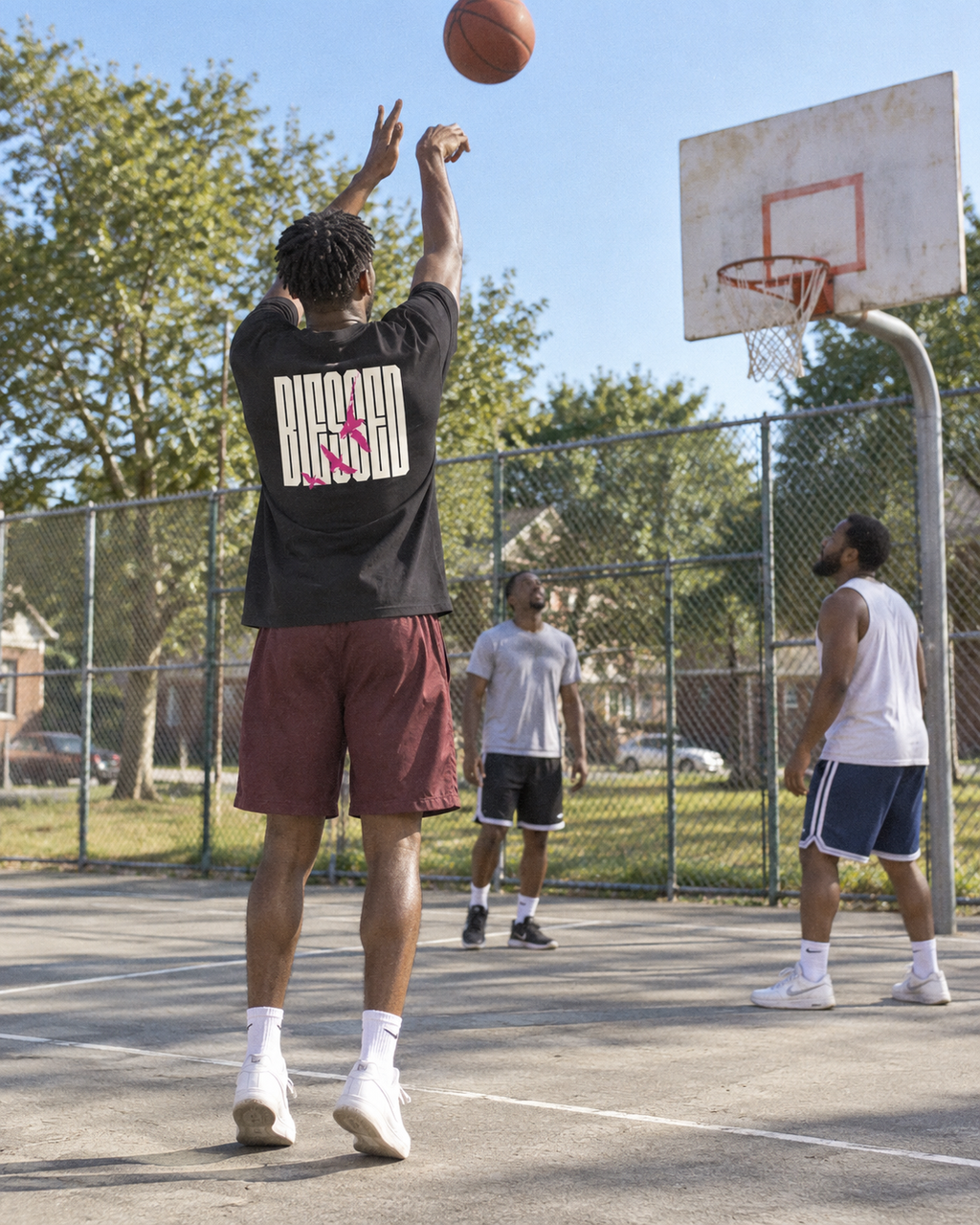 lifestyle mockup of an adult shooting a basketball on a sunny neighborhood court, seen from behind in the black Blessed Birds tee with its cream lettering and pink birds visible.