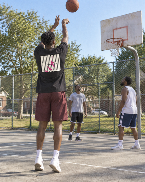 lifestyle mockup of an adult shooting a basketball on a sunny neighborhood court, seen from behind in the black Blessed Birds tee with its cream lettering and pink birds visible.
