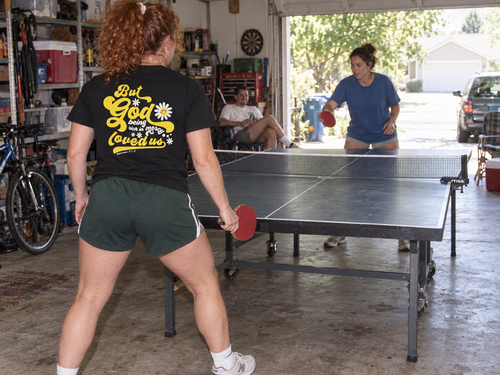 lifestyle mockup of an adult playing table tennis in the black But God Groovy tee, viewed from behind with its yellow lettering and daisy back artwork visible.