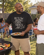 lifestyle mockup of an adult serving vegetables at a backyard cookout in the black But God Script tee, with its cream front lettering visible.