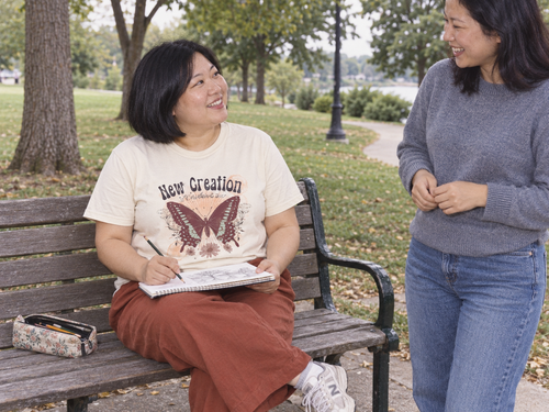 lifestyle mockup of an adult sketching on a park bench with a friend, wearing the natural Butterfly tee with its New Creation and butterfly front artwork visible.