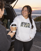 lifestyle mockup of the white Camo Jesus Is King hoodie, with front camouflage lettering, worn by a woman sharing coffee beside a vehicle at dawn.