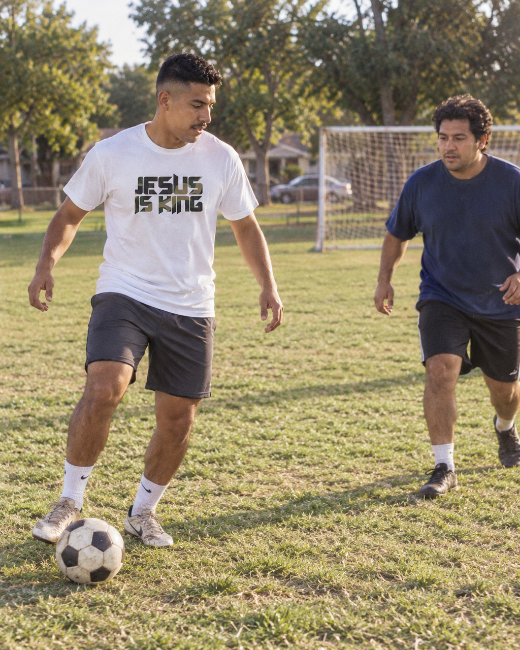 lifestyle mockup of an adult playing soccer with a friend in a park, wearing the white Camo Jesus is King tee with its camouflage front lettering visible.