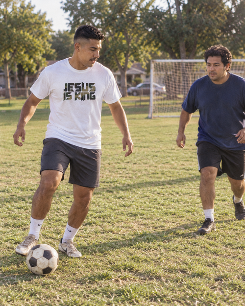 lifestyle mockup of an adult playing soccer with a friend in a park, wearing the white Camo Jesus is King tee with its camouflage front lettering visible.