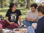 lifestyle mockup of an adult playing cards with friends on a picnic blanket, wearing the black Don’t Worry tee with its cream ship and front lettering visible.