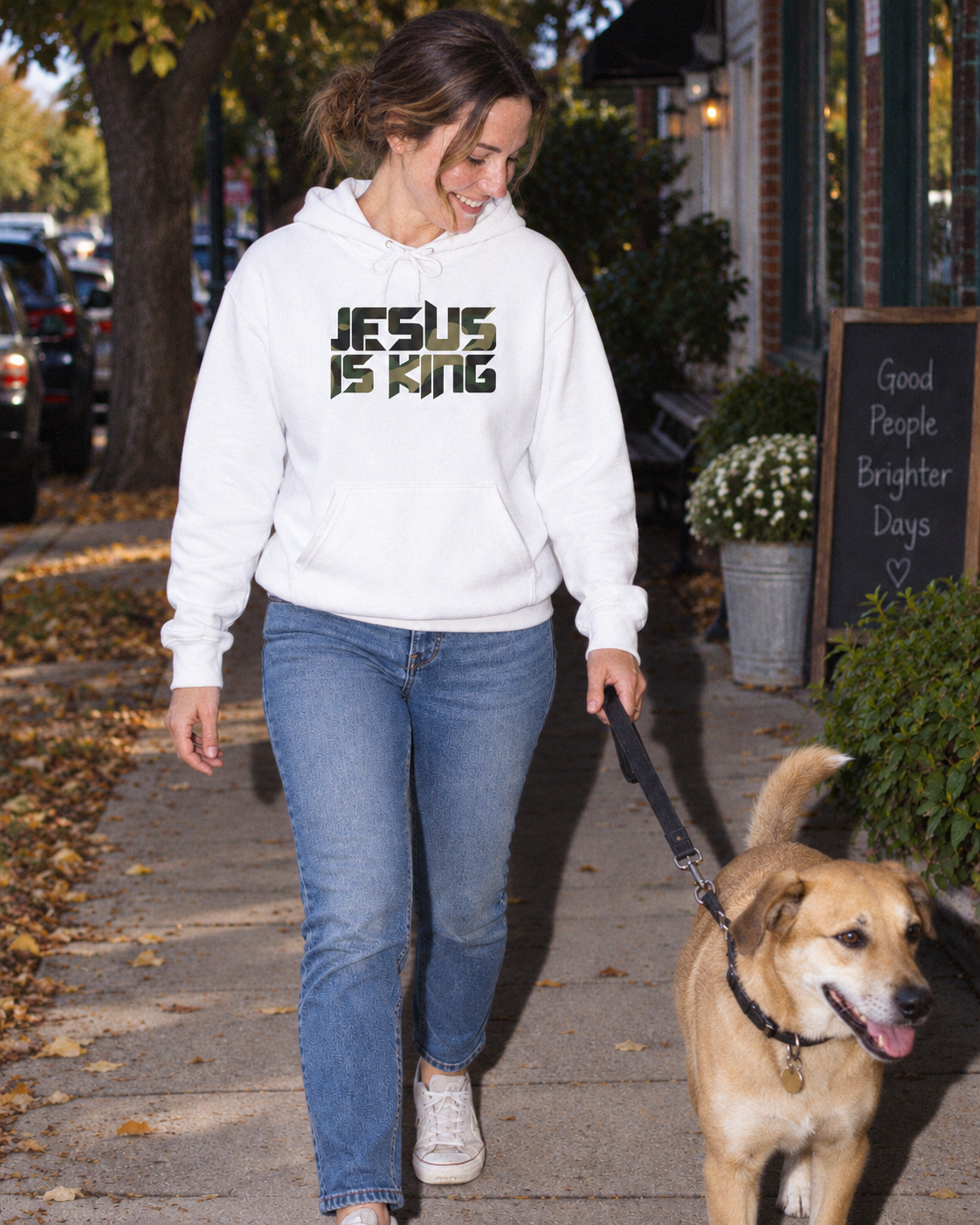 Camouflage Jesus Is King artwork on a white hoodie, worn while walking a dog on a neighborhood sidewalk