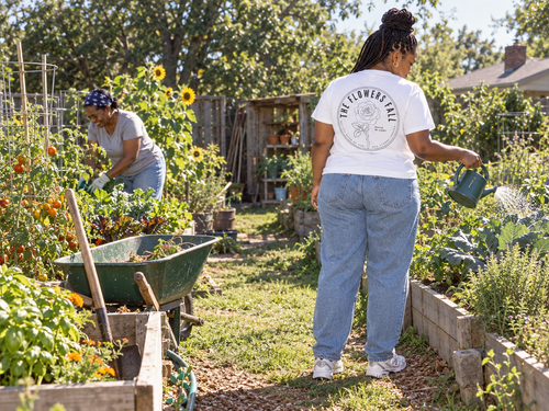 lifestyle mockup of an adult watering a raised garden bed on a sunny morning, seen from behind in the white Flowers Fall tee with its black circular rose and Isaiah 40:8 back design visible.