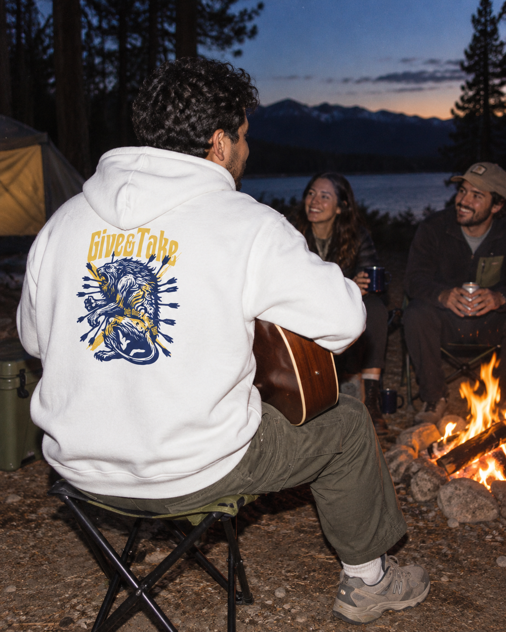 lifestyle mockup of a young adult playing guitar with friends around a campfire at dusk, seen from behind in the white Give & Take hoodie with navy and gold lion back artwork visible.