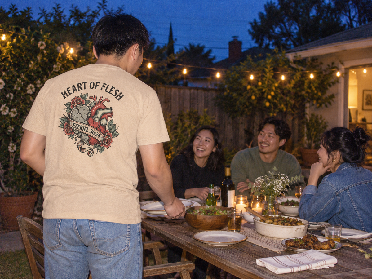 lifestyle mockup of an adult setting a backyard dinner table, seen from behind in the sand dune Heart of Flesh tee with its heart, roses and Ezekiel 36:26 back artwork.