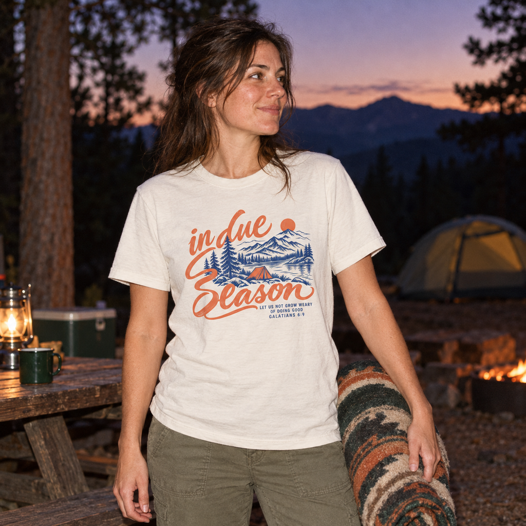 Vintage White In Due Season tee with blue and apricot mountain and camping artwork on the front, worn at a pine campground at dusk.
