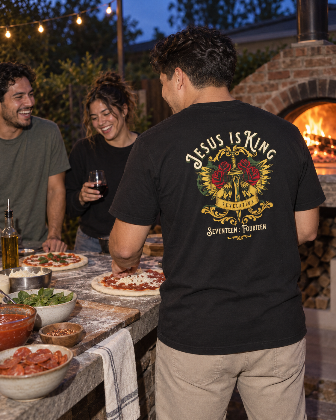 lifestyle mockup of a young adult making pizza with friends at dusk, seen from behind wearing the black Jesus Is King tee with its gold sword, red roses and cream back lettering visible.