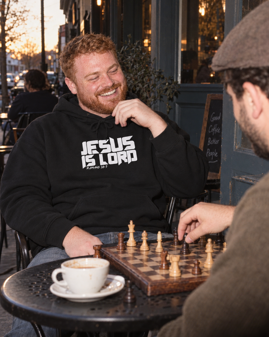 lifestyle mockup of the black Jesus Is Lord hoodie with white Romans 10:9 front artwork, worn by a man playing chess with a friend at an outdoor cafe.