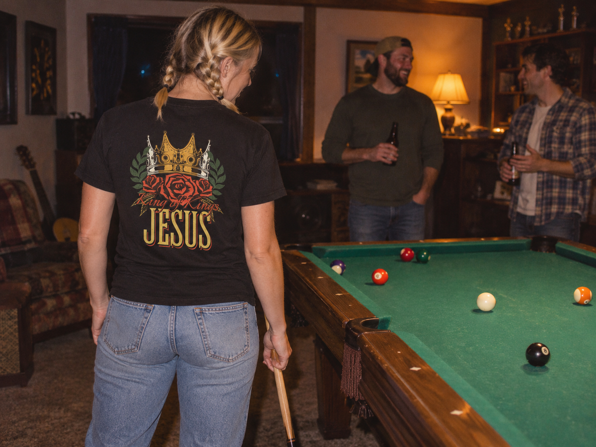 lifestyle mockup of an adult playing pool with friends in a game room, seen from behind in the black King of Kings tee with its crown and Revelation 19:16 back design visible.