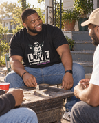 lifestyle mockup of a young adult playing dominoes with friends on a sunny patio, wearing the black Love tee with its cream cherub and LOVE front artwork visible.