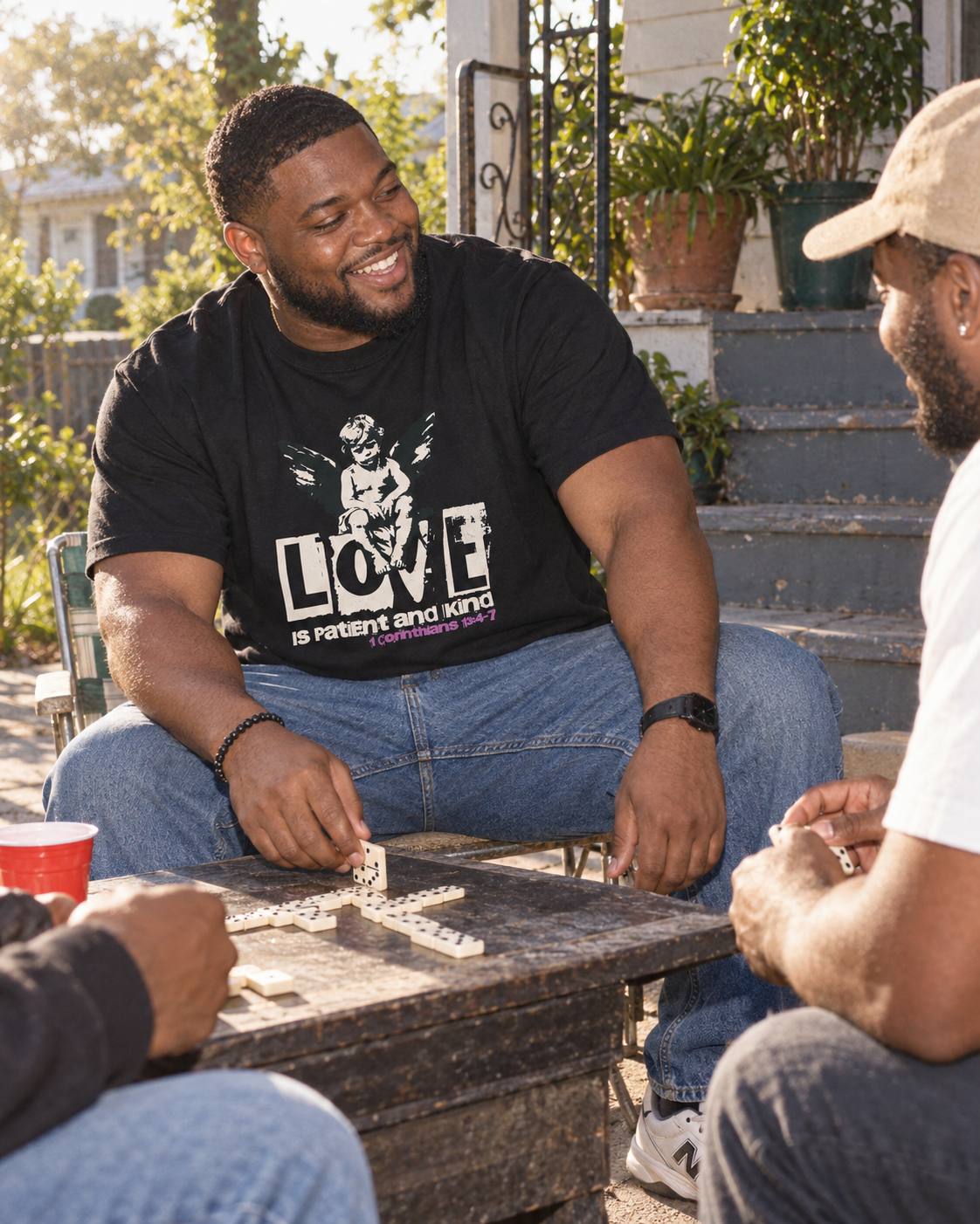 lifestyle mockup of a young adult playing dominoes with friends on a sunny patio, wearing the black Love tee with its cream cherub and LOVE front artwork visible.