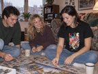 lifestyle mockup of an adult wearing the black Love II tee with its colorful floral front print, building a puzzle with friends on an apartment floor on a rainy afternoon.
