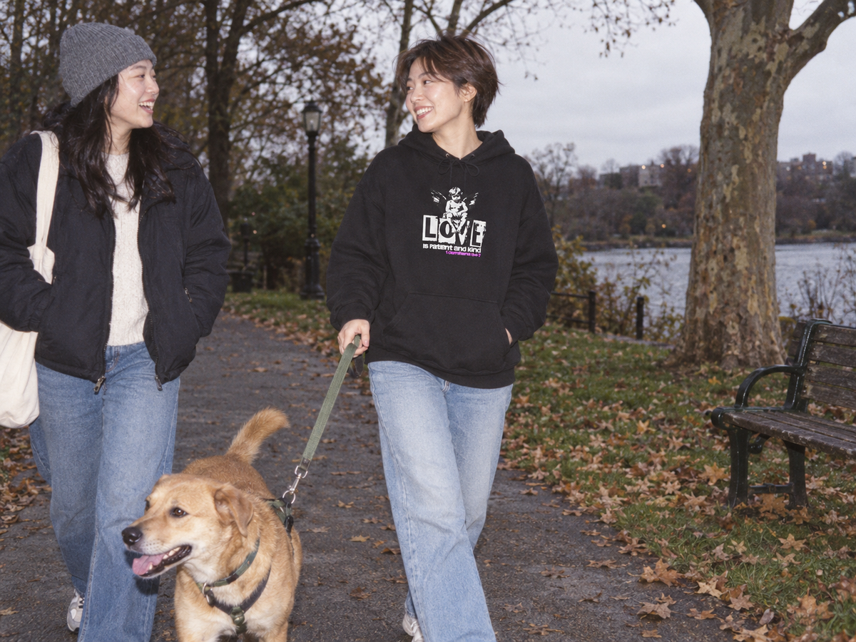 lifestyle mockup of a young adult walking a dog with a friend in a park, wearing the black Love Is Patient hoodie with its white cherub and LOVE front artwork visible.
