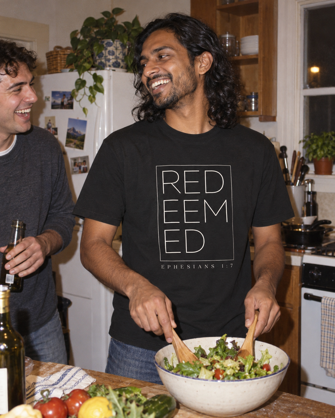 lifestyle mockup of an adult wearing the black Redeemed tee with its framed front lettering and Ephesians 1:7 print, preparing salad with a friend in a kitchen.
