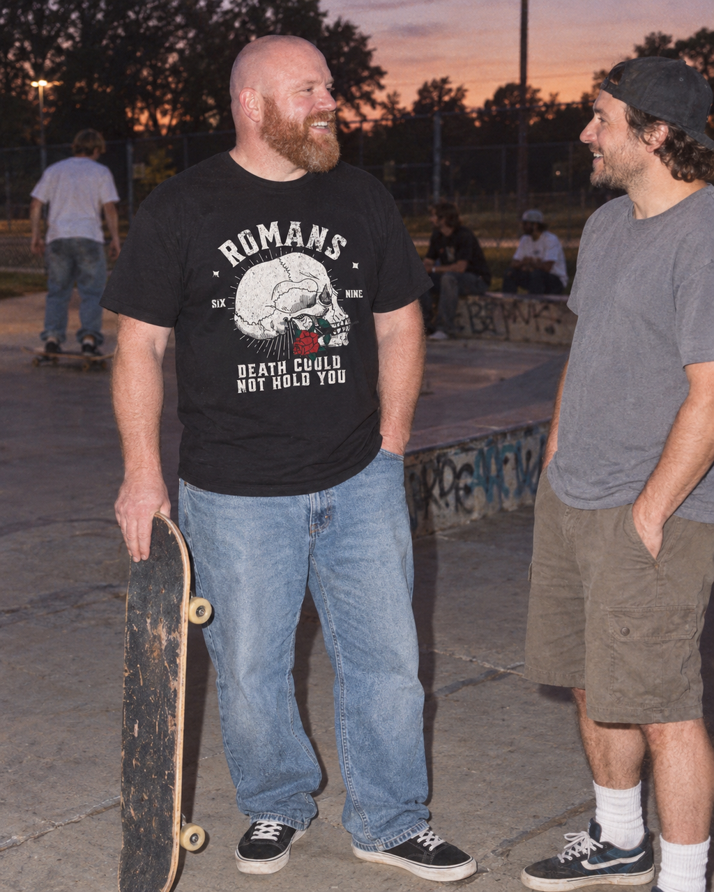 lifestyle mockup of an adult holding a skateboard while talking with a friend at dusk, wearing the black Romans Death Could Not Hold tee with its front artwork visible.