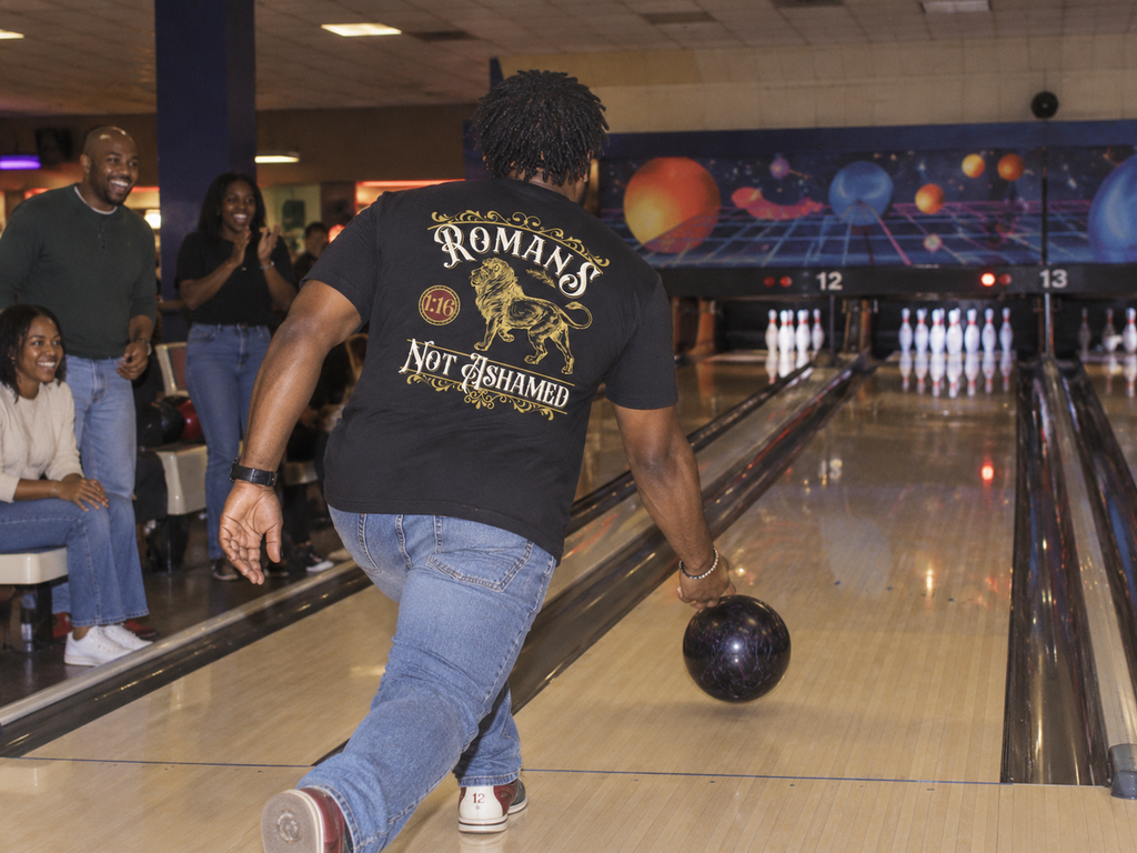 lifestyle mockup of an adult bowling with friends, photographed from behind in the black Romans Lion tee with its lion and Romans 1:16 back artwork visible.