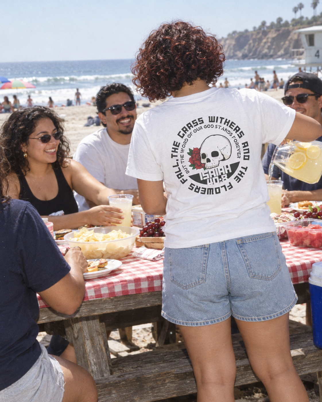 lifestyle mockup of an adult pouring lemonade for friends at a beach picnic, seen from behind in the white Rose Skull tee with its circular rose, skull and Isaiah 40:8 back artwork visible.