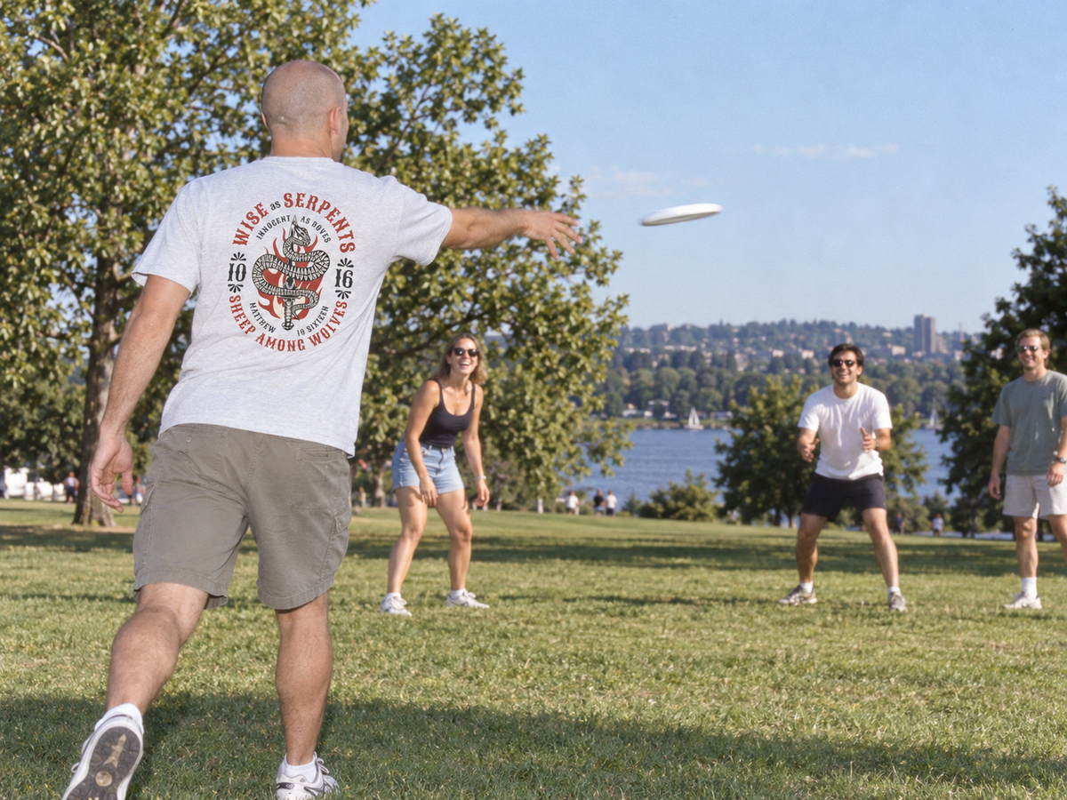 lifestyle mockup of an adult playing frisbee with friends in a sunny park, seen from behind in the ash Serpents and Doves tee with its circular back artwork visible.