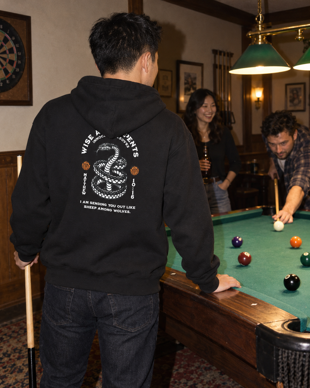 lifestyle mockup of young adult friends playing pool in the evening, with a man seen from behind in the black Serpents and Doves Classic hoodie showing its white snake, orange emblems and Matthew 10:16 back artwork.