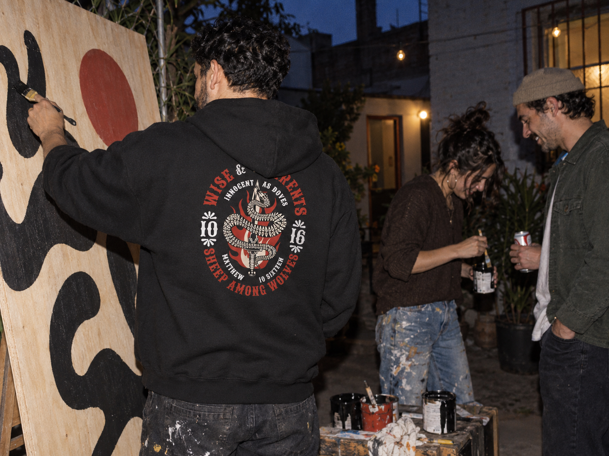 lifestyle mockup of young adult friends painting in a courtyard at dusk, seen from behind in the black Serpents and Doves hoodie with its red and cream snake-and-dagger back artwork visible.