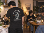 lifestyle mockup of a young adult cooking dinner with friends in an apartment kitchen, seen from behind in the black Serpents and Doves tee with its cream snake, orange emblems and Matthew 10:16 back artwork visible.
