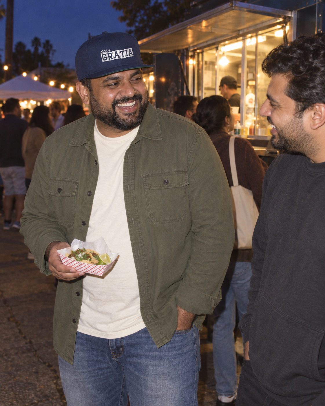 lifestyle mockup: adult wearing the navy Sola Gratia cap while waiting at a food truck with a friend at dusk; white front embroidery visible.