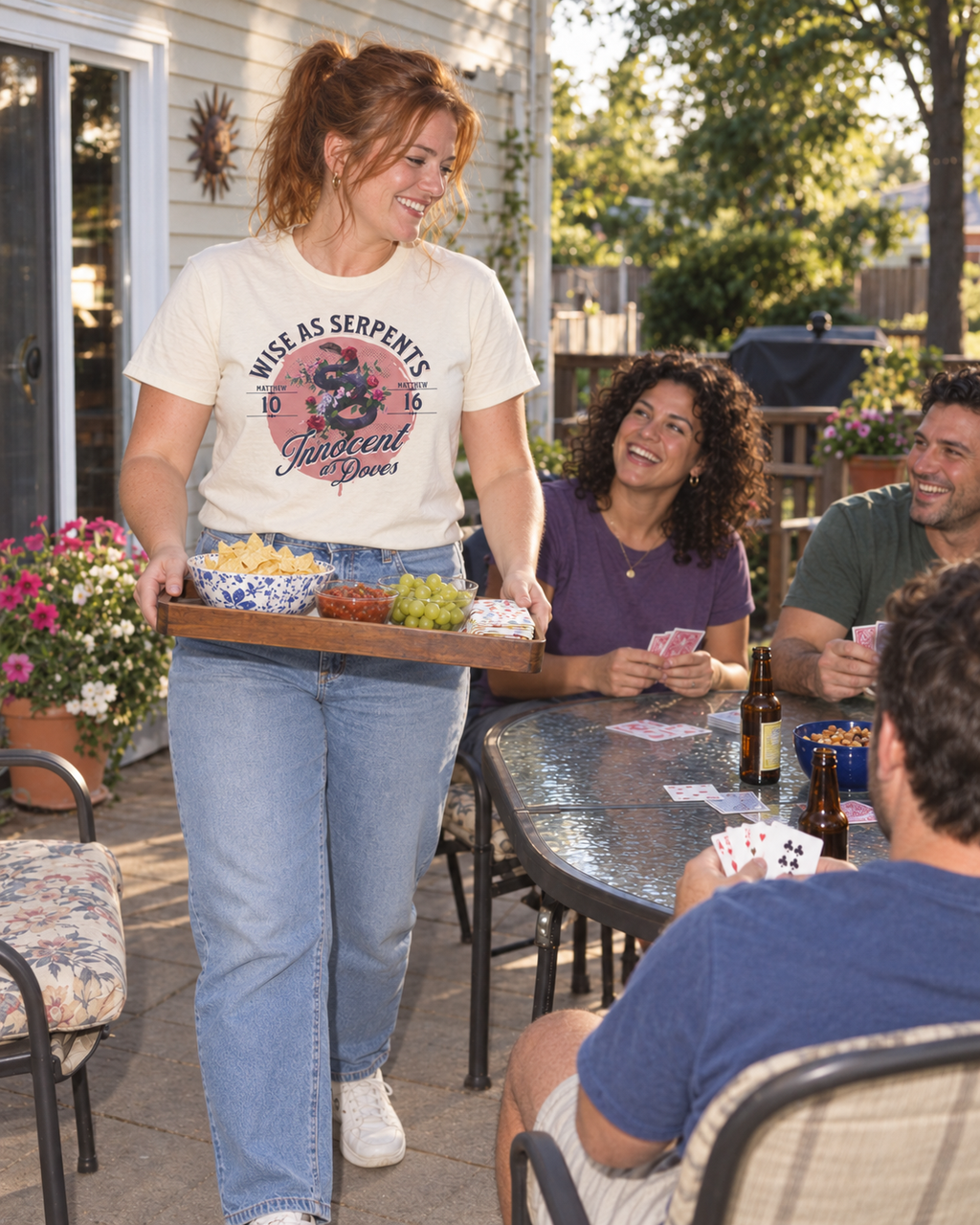lifestyle mockup of young adult friends playing cards on a sunny patio, with a woman carrying snacks in the natural Wise As Serpents Rose Snake tee and its pink rose and snake front artwork visible.