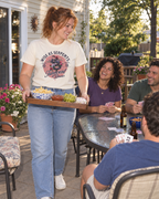 lifestyle mockup of young adult friends playing cards on a sunny patio, with a woman carrying snacks in the natural Wise As Serpents Rose Snake tee and its pink rose and snake front artwork visible.
